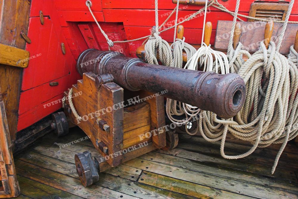 Старинное морское орудие установлено на палубе деревянного корабля, окружённое канатами.